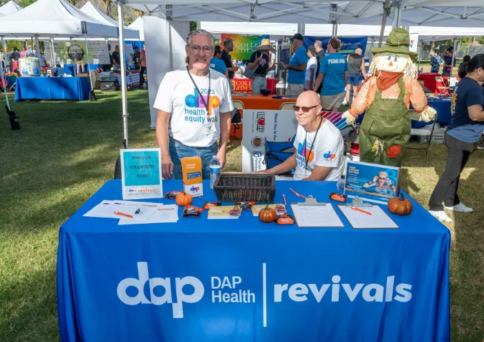 Two people sit behind a blue "DAP Health / Revivals" table booth outdoors, featuring pamphlets, small pumpkins, and artwork. Both wear branded shirts. A scarecrow decorates the display; other booths and people are visible in the background.