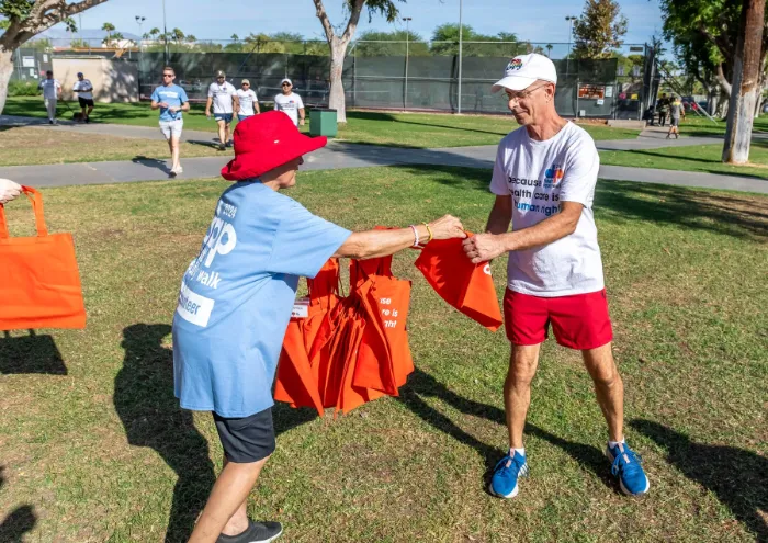 Two older adults exchange bright orange tote bags in a sunny park. One wears a red hat and blue shirt, the other a white cap, glasses, white shirt, and red shorts. Several people walk in the background near tennis courts and green grass.