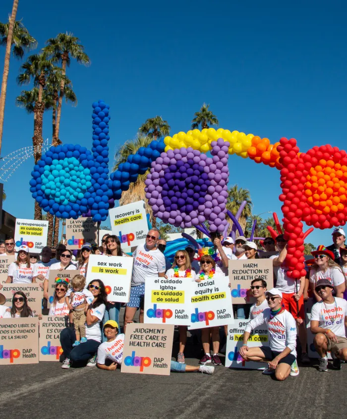 A large group of people at an outdoor pride event pose in front of a balloon sculpture spelling “dap” in rainbow colors. They hold colorful signs promoting healthcare, equality, and LGBTQ+ rights. Palm trees and a clear blue sky are in the background.
