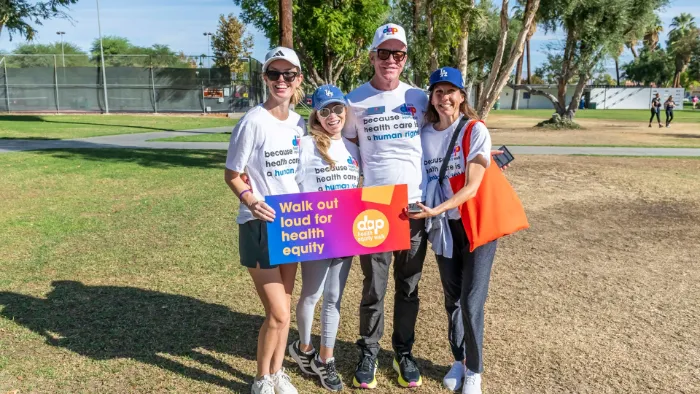 Four people stand outdoors on grass, smiling at the camera. They wear white shirts with health care slogans and hold a sign reading “Walk out loud for health equity.” Trees and a tennis court are visible in the background on a sunny day.