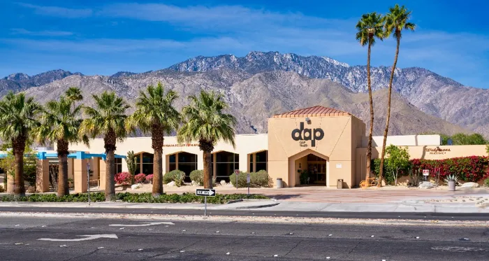 A tan building with “dap” written on the front stands amid palm trees, with mountains rising in the background under a blue sky. The scene is sunlit, with landscaped bushes and flowers around the entrance and a wide street in the foreground.