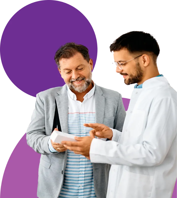 Two men talk together. One, with gray hair and a beard, wears a light gray blazer and smiles while holding a notepad. The other, younger with short dark hair and glasses, wears a white lab coat and gestures at the notepad. Purple graphics are in the background.