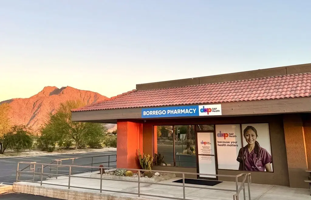 A pharmacy with a red tile roof and sign reading "Borrego Pharmacy" is pictured at sunset. A poster on the window shows a smiling healthcare worker. Mountains, trees, and desert vegetation are visible in the background under a clear sky.