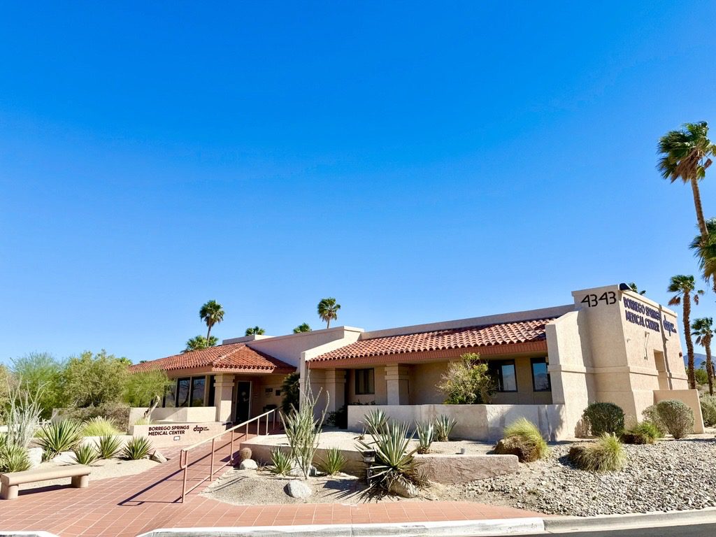 A beige, southwestern-style medical clinic with a red-tiled roof and desert landscaping sits under a clear blue sky. The sign reads "PRIMARY CARE DOCTOR" and the address "4343" is visible on the building. Palm trees are scattered in the background.