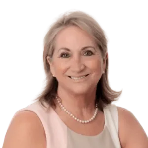 A smiling older woman with light skin and shoulder-length blonde hair wears a sleeveless light pink top, a pearl necklace, and light makeup. She is facing the camera, photographed against a plain white background.
