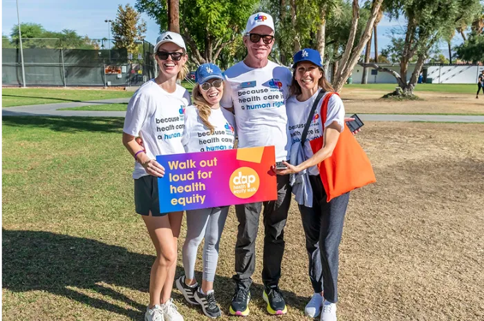 Four smiling people stand outdoors on grass, wearing casual clothes and hats. Two wear shirts reading “because health care is a human right.” They hold a colorful sign: “Walk out loud for health equity.” Trees, a tennis court, and blue sky are in the background.