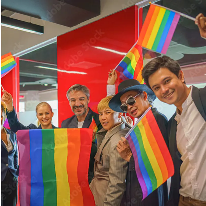 A diverse group of smiling people in business attire stand indoors, holding rainbow pride flags. They pose together in front of a red wall, celebrating LGBTQ+ pride in a modern office setting.