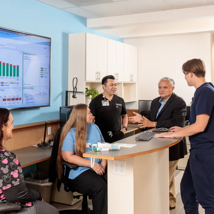 Five people, including medical staff in scrubs, are having a discussion around a curved workstation in a clinic. A large screen displays charts and data. Cabinets, monitors, and medical equipment are visible in the bright, modern room.