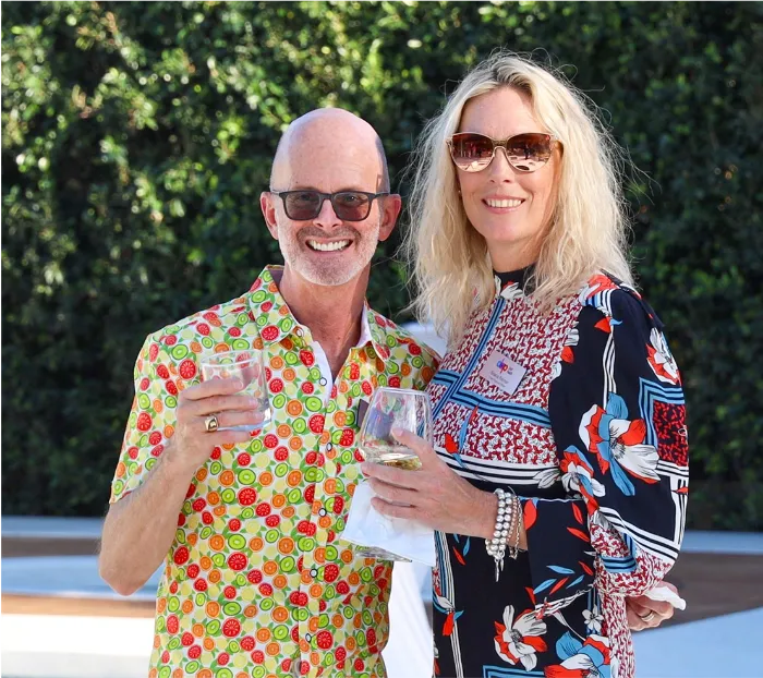 A smiling man in a colorful shirt and glasses stands next to a smiling woman in a patterned dress and sunglasses. Both hold drinks at a lively DAP Health Events and Fundraisers gathering, enjoying the green foliage in the background.