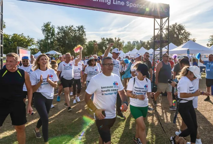 A diverse group of people, many wearing white shirts that read “because health care is a human right,” are walking outdoors under a finish line banner at a sunny event; some are smiling, and tents and trees are visible in the background.
