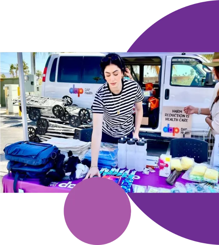 A person arranges supplies, including water bottles and hygiene items, on a purple table outside a DAP Health van. Another person stands nearby. The van and table are branded with DAP Health logos. The scene is outdoors on a sunny day.
