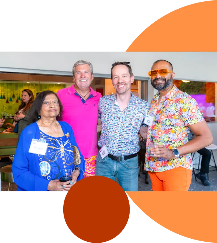 Four people stand smiling at an indoor gathering. The group includes an older woman in a blue patterned blouse and three men in colorful shirts. One man wears orange sunglasses and pants. They each wear name tags and hold drinks, with a festive background behind them.