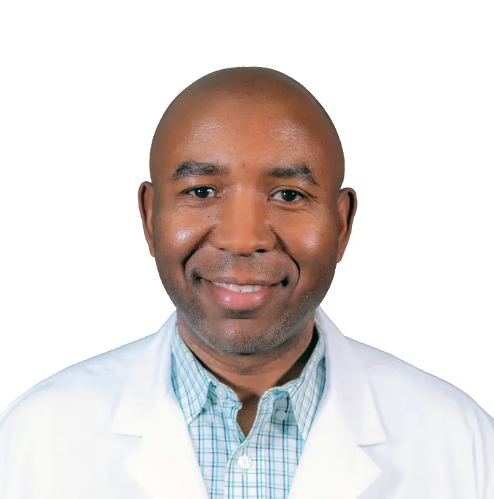 A man with a shaved head, wearing a white lab coat over a light plaid shirt, smiles at the camera. The background is plain white, and he appears professional and approachable, likely in a medical or scientific field.