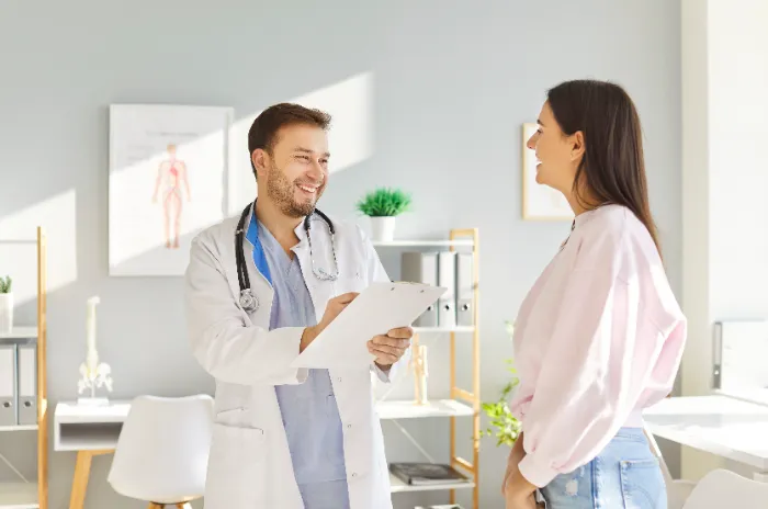 A smiling male doctor in a white coat and stethoscope holds a clipboard and talks with a woman in a light pink sweater in a bright, modern medical office. Both appear happy and engaged. Shelves, plants, and medical posters are visible in the background.