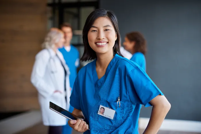 A smiling healthcare worker in blue scrubs holds a tablet and stands in focus. Her ID badge with a photo is clipped to her top. In the blurred background, three other medical professionals in white and blue coats talk together.