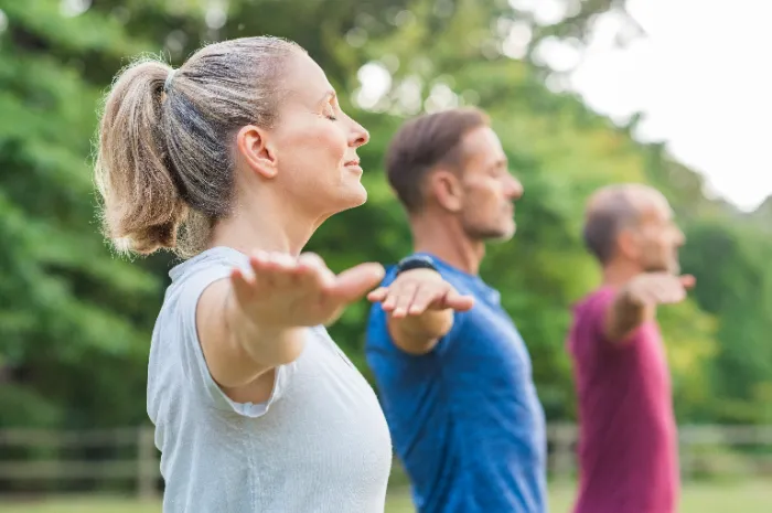 Three adults stand outdoors with arms extended at shoulder height, eyes closed, and faces relaxed, practicing a mindfulness or stretching exercise. They are positioned side by side, facing left, with lush green trees in the blurred background.