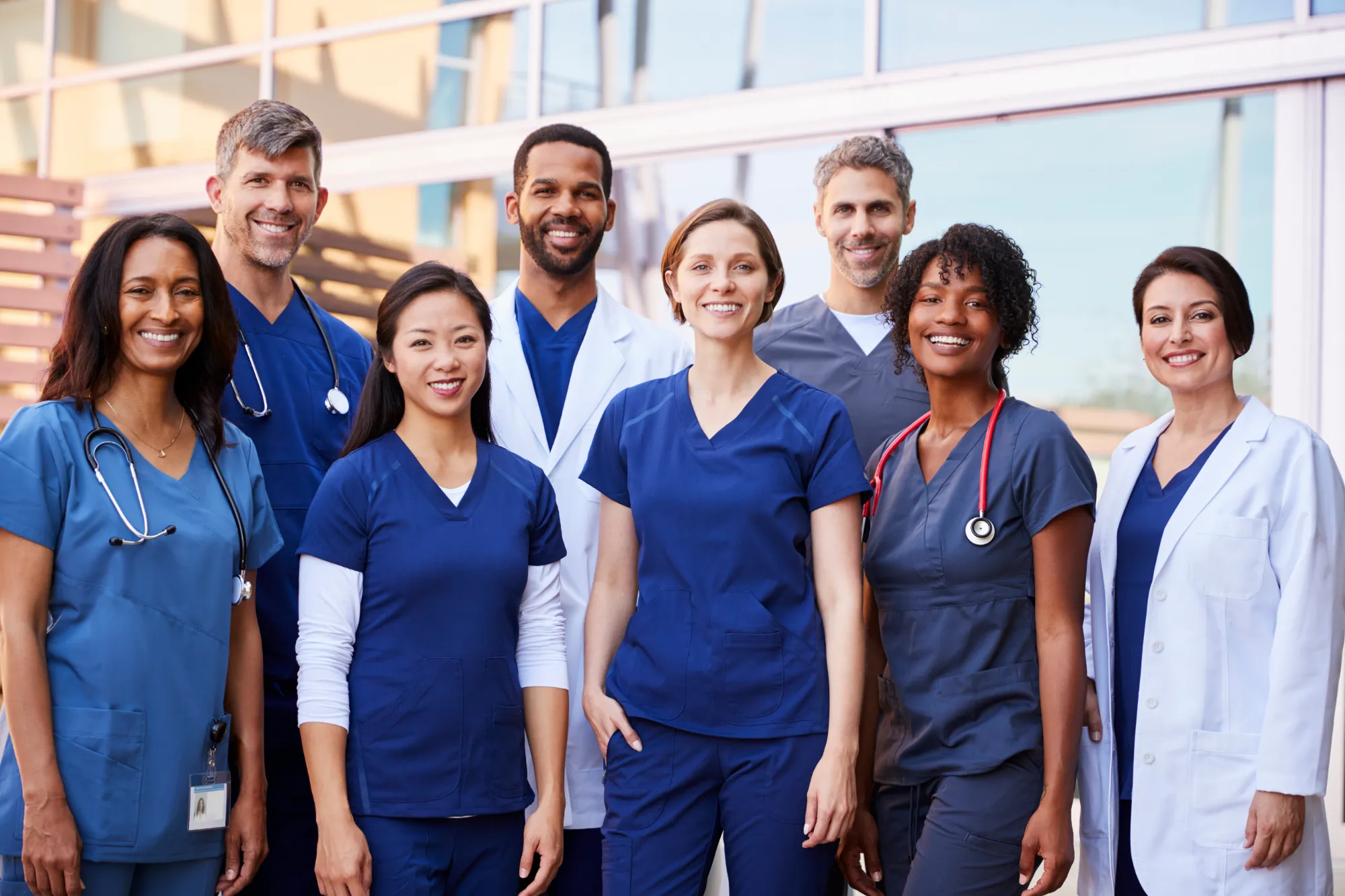 A diverse group of eight smiling healthcare professionals, wearing scrubs and lab coats, stand together outdoors in front of a modern building, suggesting teamwork and unity in a medical or hospital setting.