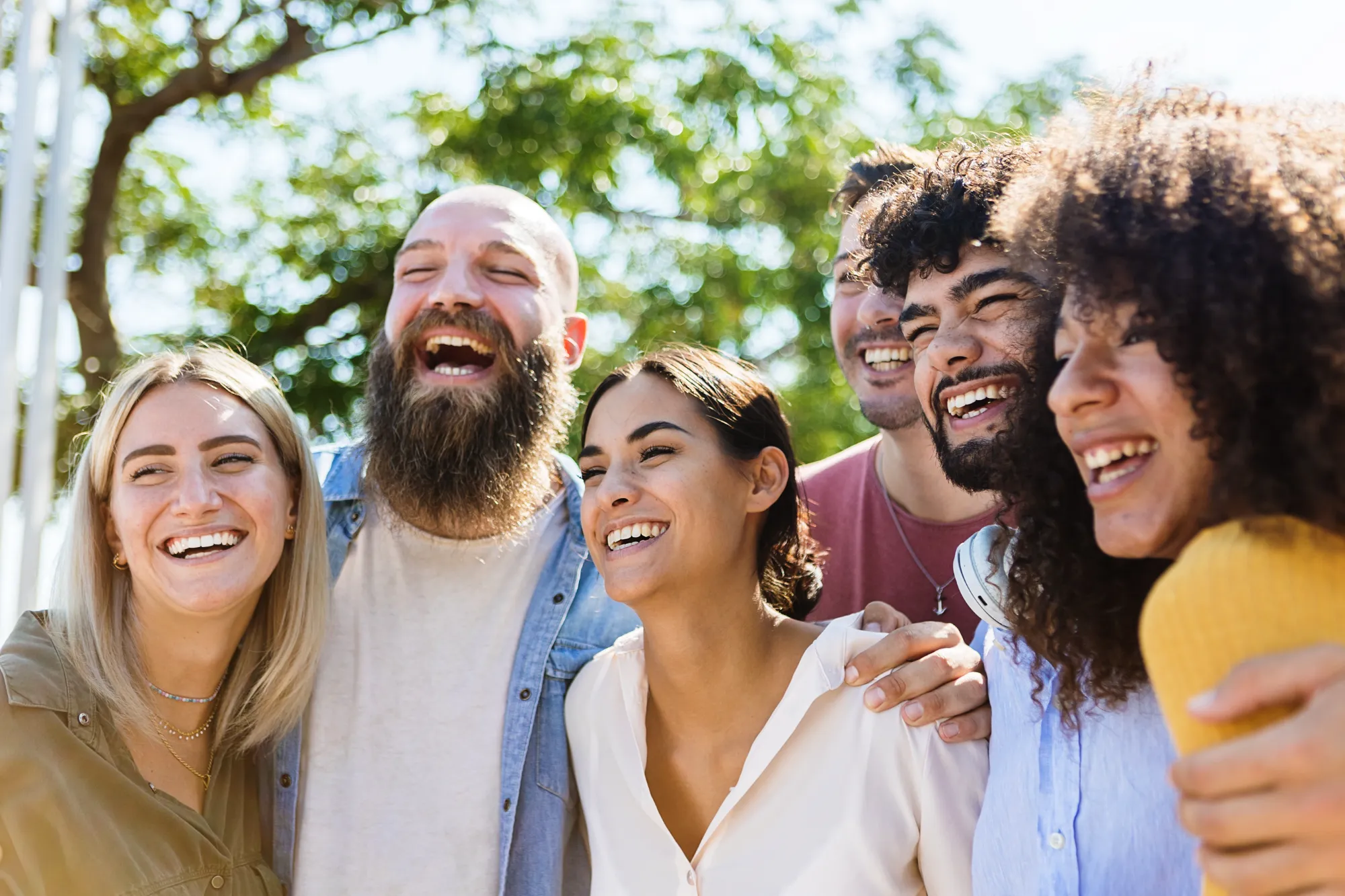 A diverse group of six young adults stands close together outdoors, smiling and laughing. They appear joyful and friendly, with trees and sunlight in the background, suggesting a warm, happy atmosphere on a bright day.