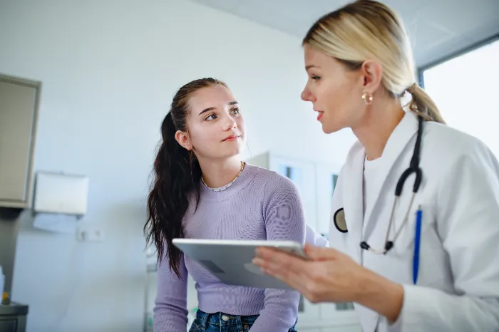 A female doctor in a white coat holds a tablet and talks to a teenage girl with long brown hair, who is listening attentively. They are in a bright medical office with cabinets and medical equipment in the background.