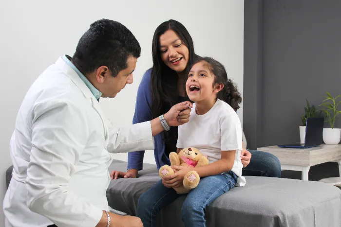 A doctor examines a smiling young girl’s throat as she sits on an exam table holding a teddy bear. A woman, possibly her mother, sits beside her, smiling. The room is bright and modern, with a desk and plants in the background.