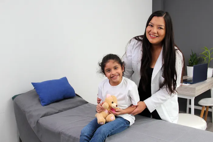 A smiling woman in a white coat, likely a doctor, sits beside a smiling young girl holding a teddy bear. They are in a medical examination room with a bed, gray bedding, a blue pillow, and a desk with a plant and laptop in the background.