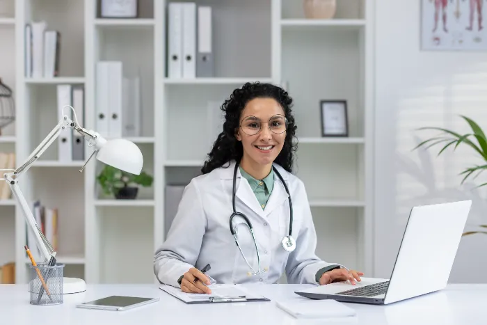 A smiling doctor with curly hair, wearing glasses, a white coat, and a stethoscope, sits at a desk with an open laptop and clipboard in a bright office. Shelves with books and plants are in the background.