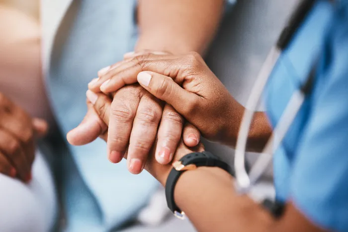 A close-up of a healthcare worker wearing blue scrubs and a stethoscope gently holding the hands of a patient, conveying comfort and support. The patient’s hand rests on top, while the worker’s hands are underneath, offering reassurance.