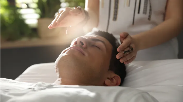 A man lies on his back with eyes closed, appearing relaxed, while a person holds their hands hovering above his face, as if performing energy healing. Soft sunlight filters in, creating a peaceful atmosphere. Green plants are blurred in the background.