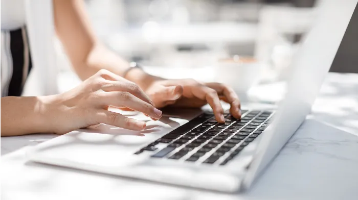 Close-up of a person’s hands typing on a laptop keyboard at a bright, white table. The background is softly blurred, with a cup and other indistinct objects visible, suggesting a casual or work setting. The lighting is natural and well-lit.