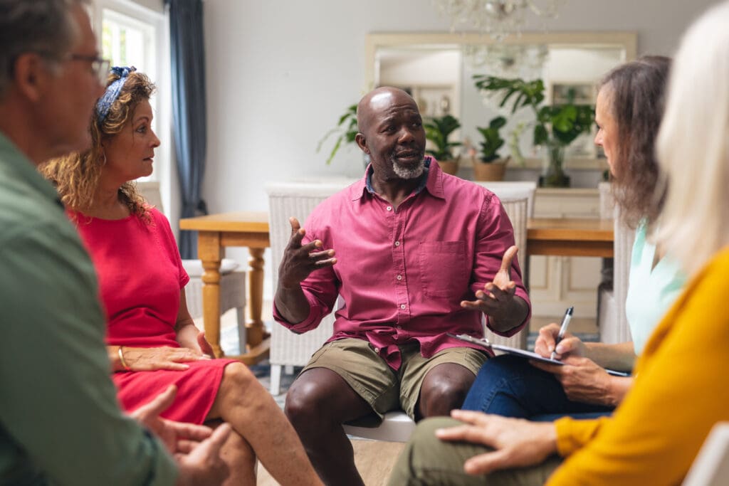A group of five adults sits in a bright living room, engaged in conversation. The man in the center, wearing a pink shirt and shorts, gestures while speaking. Others listen attentively, some taking notes. Green plants and a large mirror decorate the background.