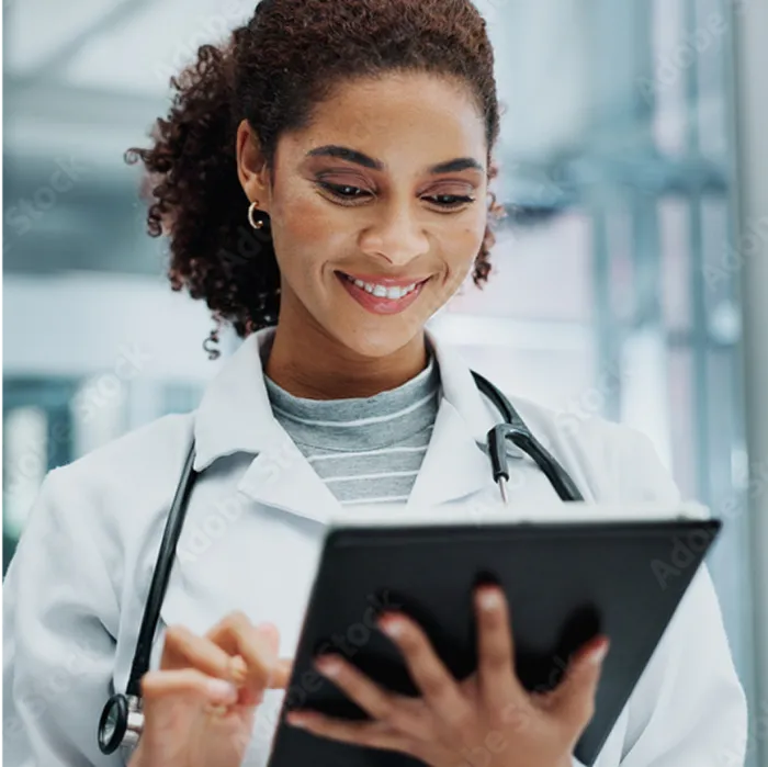 A woman in a white lab coat with a stethoscope around her neck smiles while using a digital tablet. She has curly hair pulled back and is standing in a bright, modern medical setting with blurred background elements.