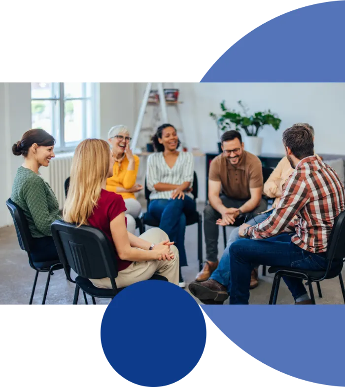 A diverse group of adults sit in a circle on chairs in a bright room, smiling and talking. The atmosphere appears relaxed and friendly. Large blue geometric shapes partially frame the photo’s edges.