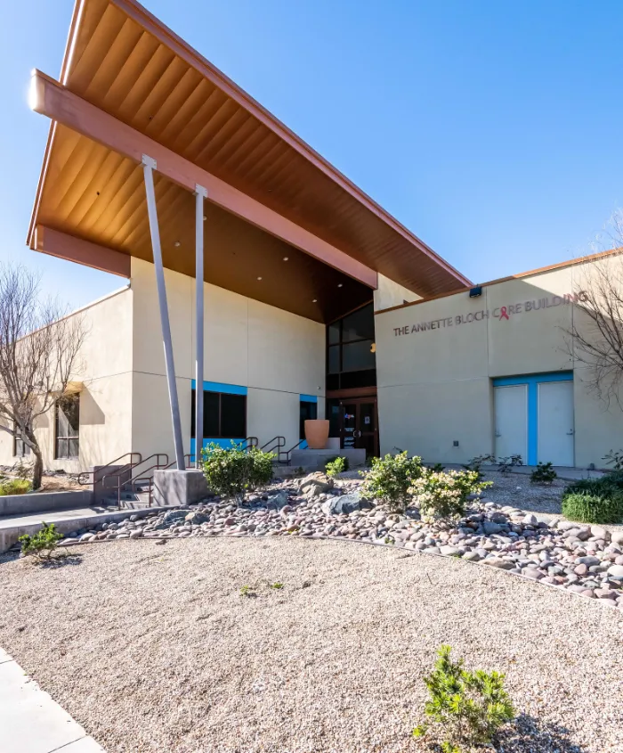 Modern beige building with angular roof and tall metal supports, labeled "The Annette Bloch Care Building." Large windows, blue sky, landscaped entry with rocks, bushes, and some leafless trees, create a clean and welcoming appearance.