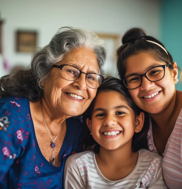 Three generations of women smile closely together. An elderly woman with gray hair and glasses is on the left, a young girl with long dark hair is in the middle, and a woman with glasses and a headband is on the right. All are happy and affectionate.