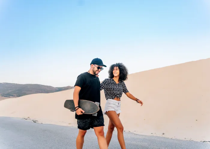 A smiling man in black clothes and sunglasses walks beside a woman in a patterned top and white shorts on a road by sandy dunes. The man holds a skateboard, and they appear happy, enjoying a sunny, clear day. Hills are visible in the distant background.