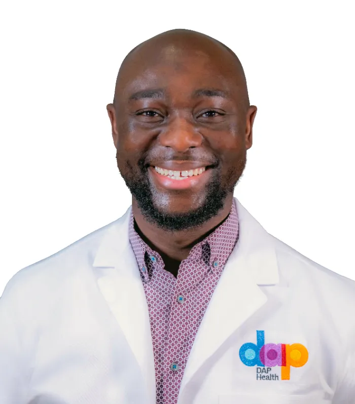A smiling man with a bald head and beard wears a white lab coat with the colorful "dap Health" logo over a pink patterned shirt. The background is plain white.