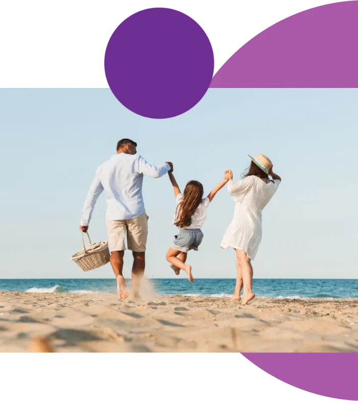 A man, woman, and young girl walk together on a sandy beach. The man holds a picnic basket and the woman wears a sunhat. Both adults lift the girl by her hands. The sea and blue sky are in the background, with purple geometric shapes overlaying the image.