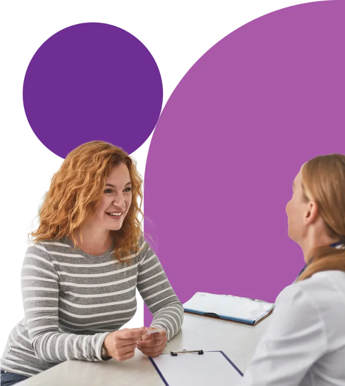 Two women sit at a table having a conversation. One, with curly red hair and a striped shirt, smiles while holding a card. The other, seen from behind, wears a white coat. A clipboard and pen are on the table. Purple circles decorate the background.