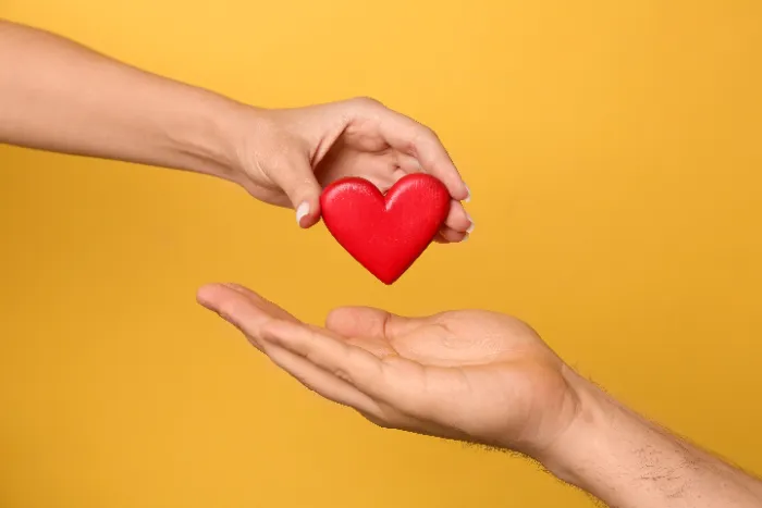 A close-up of two hands against a solid yellow background. One hand is holding a red heart-shaped object and lowering it into the open palm of another hand below, symbolizing giving, love, care, or support.