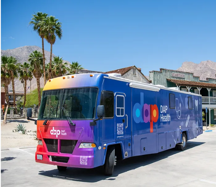 A large blue and purple DAP Health mobile clinic is parked on a sunny street with palm trees and desert mountains in the background. The bus features logos, a stethoscope graphic, and a QR code, with rustic wooden buildings behind it.