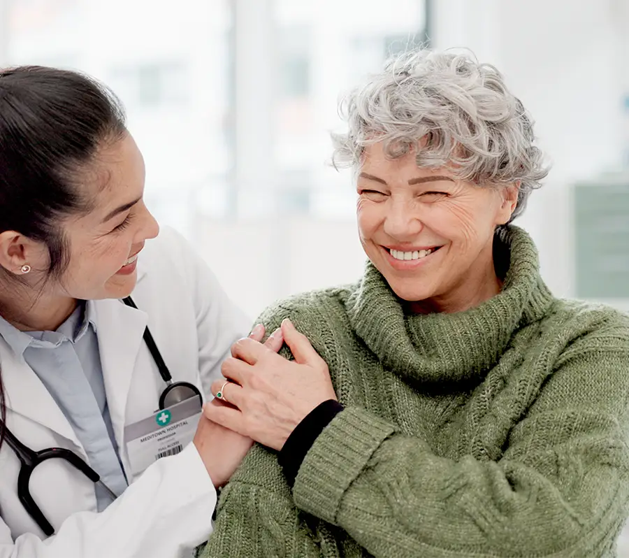 A smiling older woman with curly gray hair and a green sweater sits while a female doctor holds her arm comfortingly. The doctor, wearing a white coat and stethoscope, smiles warmly, conveying care and support in a bright, clinical setting.
