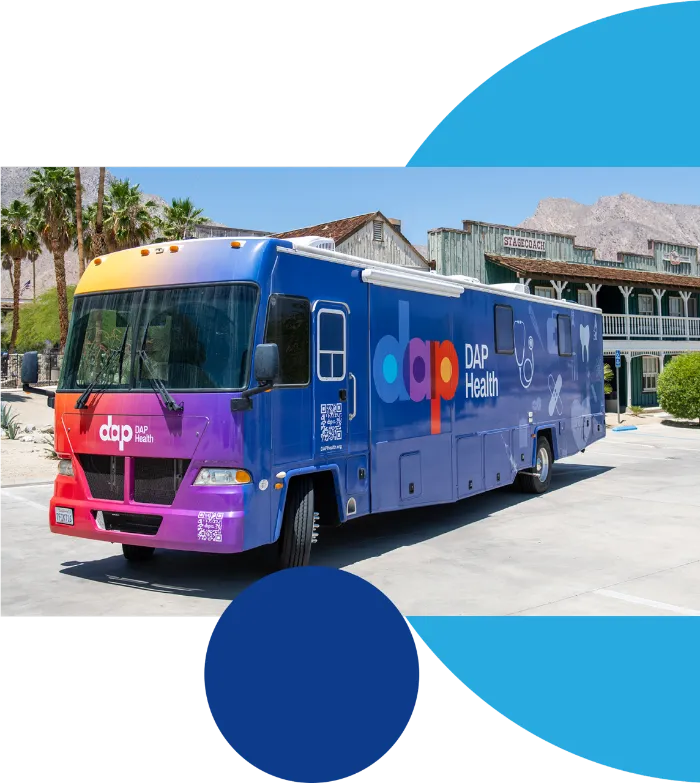 A colorful DAP Health mobile clinic RV is parked outside, featuring a vibrant gradient design and DAP Health logo. Palm trees, a historic wooden building, and mountains are visible in the sunny background.