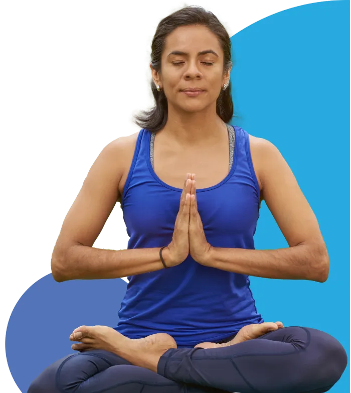 A woman sits cross-legged in a yoga pose with her eyes closed and hands pressed together in prayer position. She wears a blue tank top and dark leggings, meditating calmly against a blue abstract background.