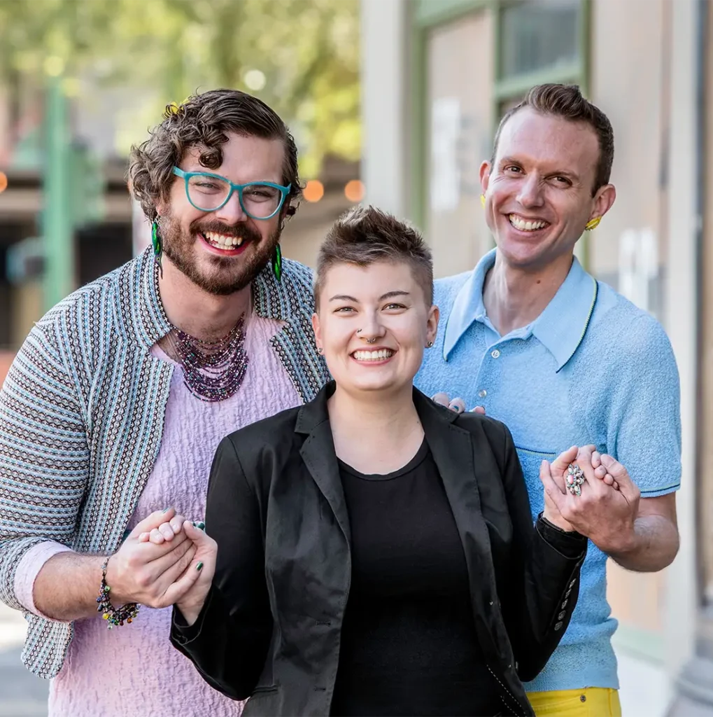 Three people stand outdoors, smiling and holding hands. Two wear colorful outfits and accessories, including earrings and glasses; the third, in the middle, wears a black blazer. The background is blurred, showing greenery and buildings.