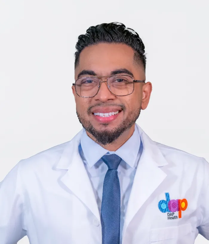 A smiling person with short, dark, styled hair, trimmed beard, and glasses wears a white DAP Health lab coat over a blue shirt and tie, standing against a plain light background. The colorful DAP Health logo is visible on the coat.