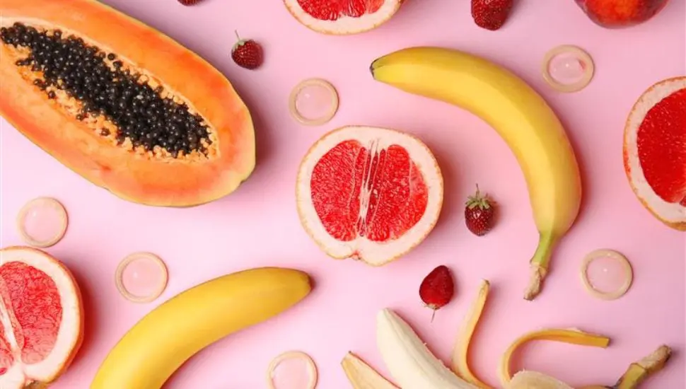 A flat lay of halved papaya, grapefruits, bananas (whole and peeled), strawberries, and unwrapped condoms on a pink background, arranged in a playful, colorful pattern.