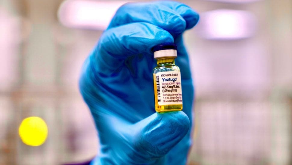 A gloved hand holds up a small vial labeled “Vaxcyte” with yellow liquid inside. The background is out of focus, suggesting a medical or laboratory setting. The blue glove contrasts with the vial’s label and contents.