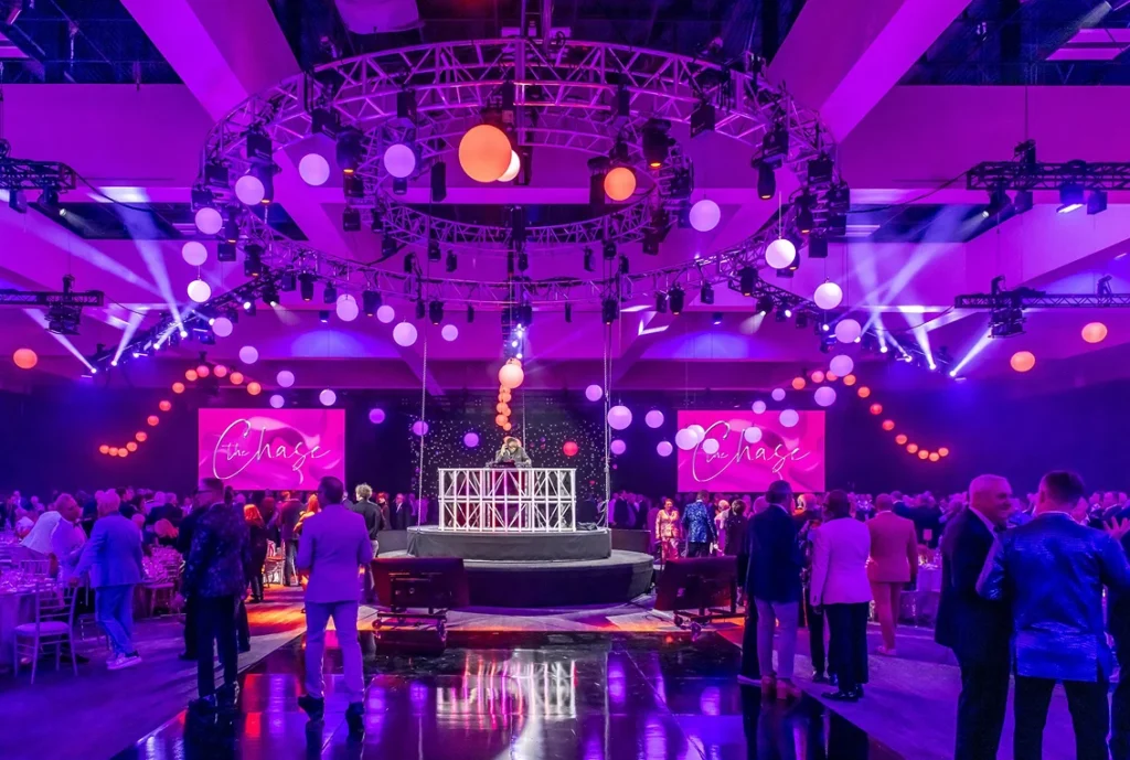A vibrant indoor event features a crowd of people in formal attire, colorful lighting, and hanging spherical lights. A stage with a white circular railing stands center, with large screens displaying “Change” in script surrounded by pink-purple lights.