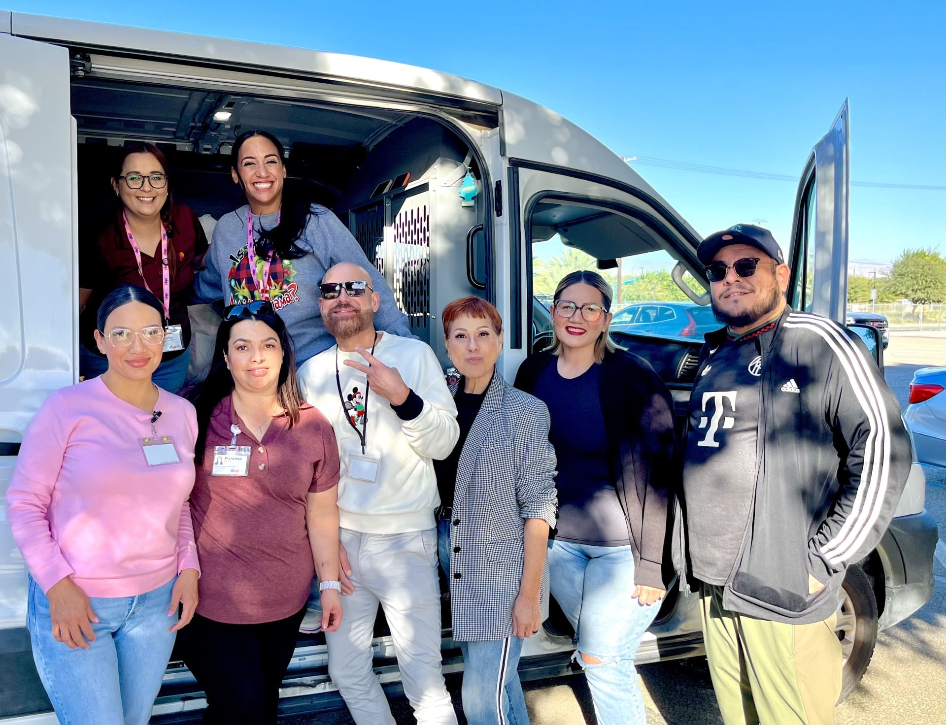 Eight adults smile and pose together outside a white van on a sunny day. Some stand, while others sit in the van doorway. They wear casual clothing and badges, suggesting work or volunteering, with a clear blue sky and trees in the background.