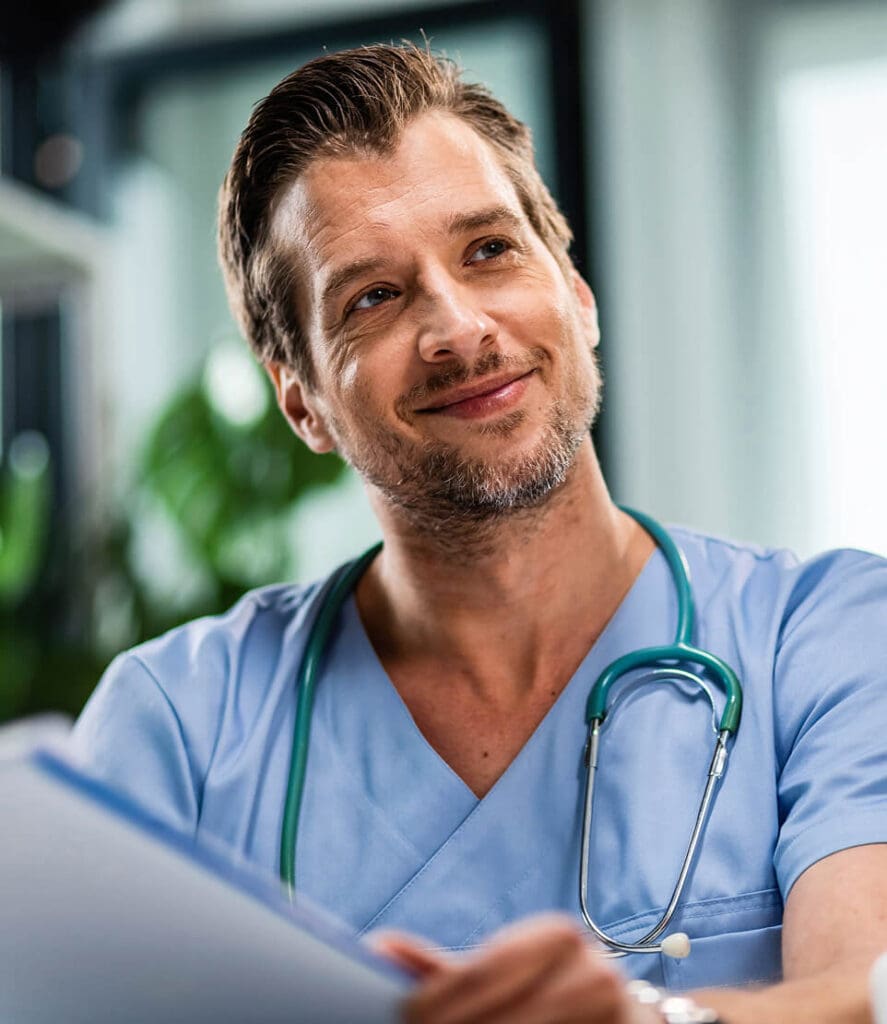 A smiling man in blue medical scrubs with a green stethoscope around his neck holds a clipboard. He has short, neatly styled brown hair and some facial stubble. The background is blurred, suggesting a medical or office setting.